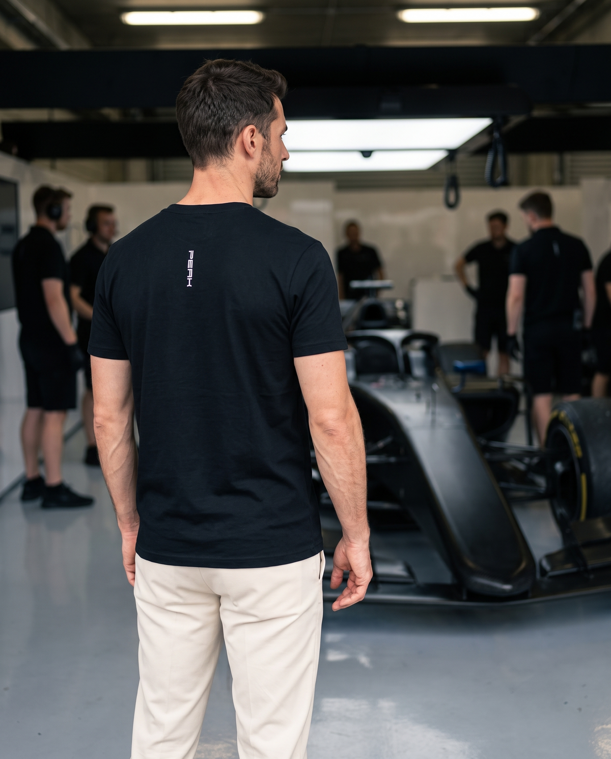 Back view of the PEAK Tee, Chest & Spine Print showing the vertical spine wordmark in a race team garage