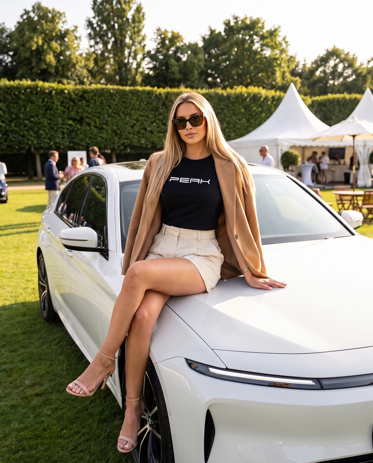 Woman wearing the PEAK Women's Maple Tee in Black with a camel blazer, seated on a white car at a grand prix garden