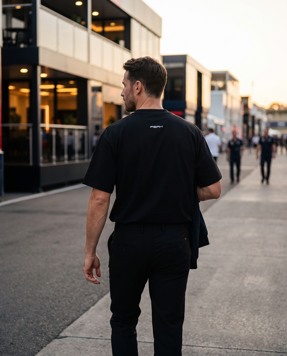 PEAK Back Neck Tee in Black worn walking through a race paddock at dusk