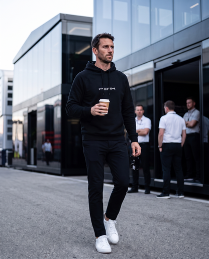 Man wearing the PEAK Supply Hoodie in Black walking through a race paddock with a coffee