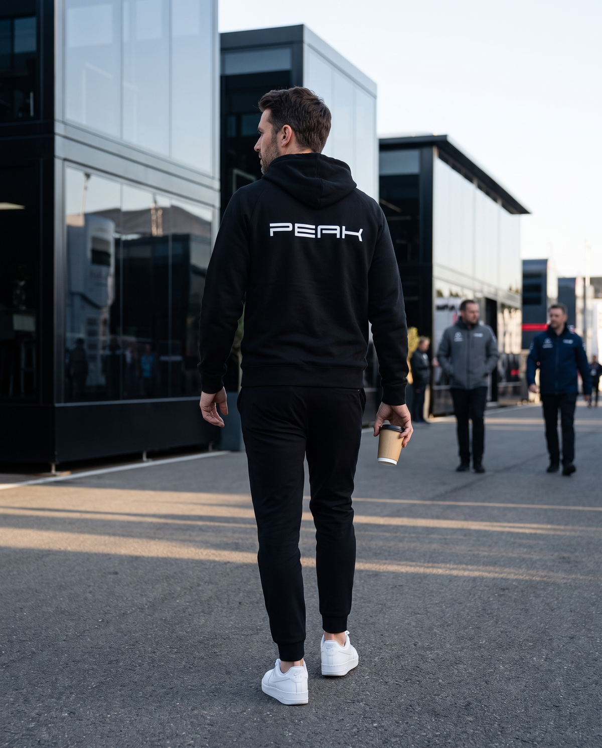 Back view of the PEAK Supply Hoodie in Black with the back print, walking through a race paddock in the morning