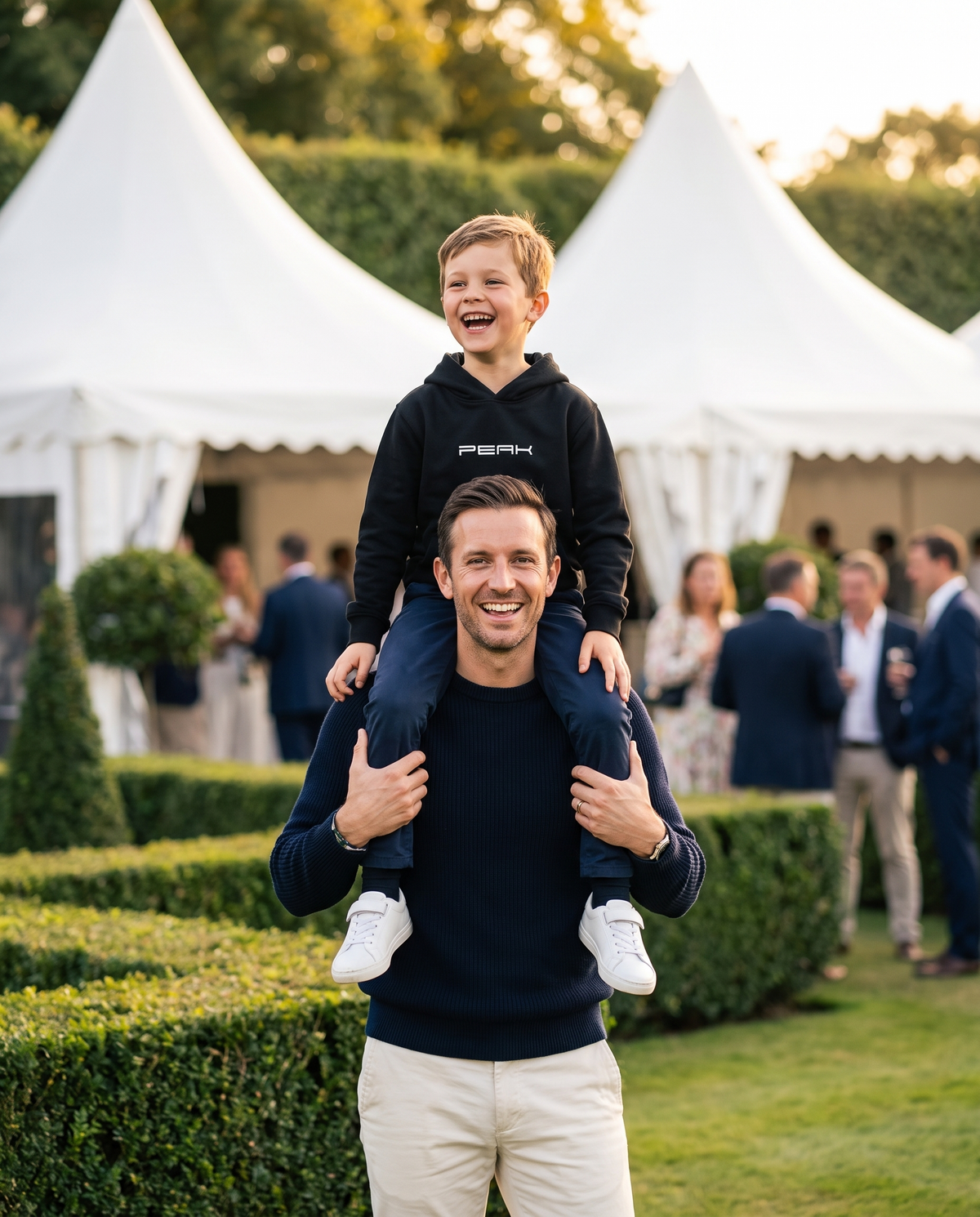 Boy in the PEAK Kids Hoodie in Black on his dad's shoulders at a grand prix hospitality garden